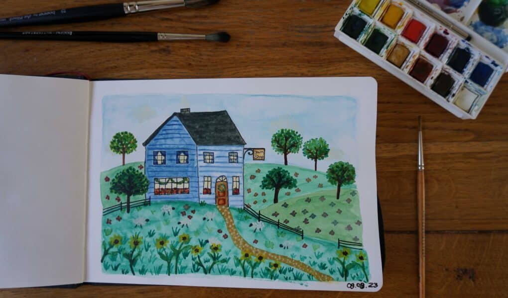 open sketchbook with watercolor and ink painting of a blue yarn shop in the hills