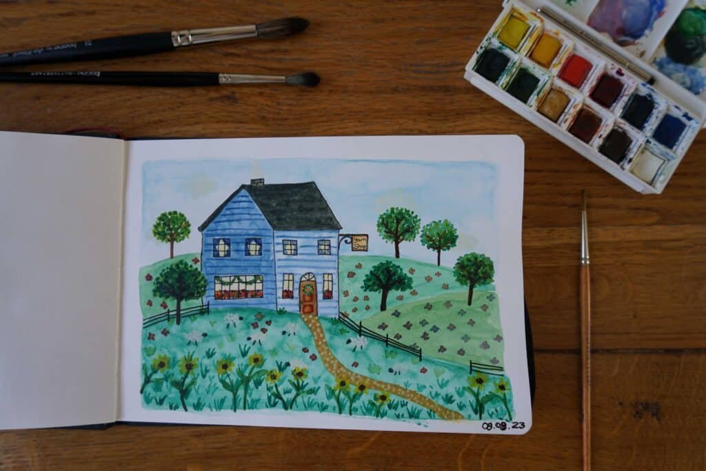 open sketchbook with watercolor and ink painting of a blue yarn shop in the hills