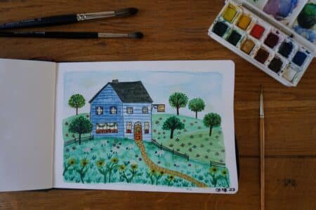 open sketchbook with watercolor and ink painting of a blue yarn shop in the hills