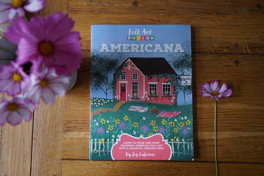 book folk art fusion americana by joy laforme on a wooden table next to some light pink cosmos