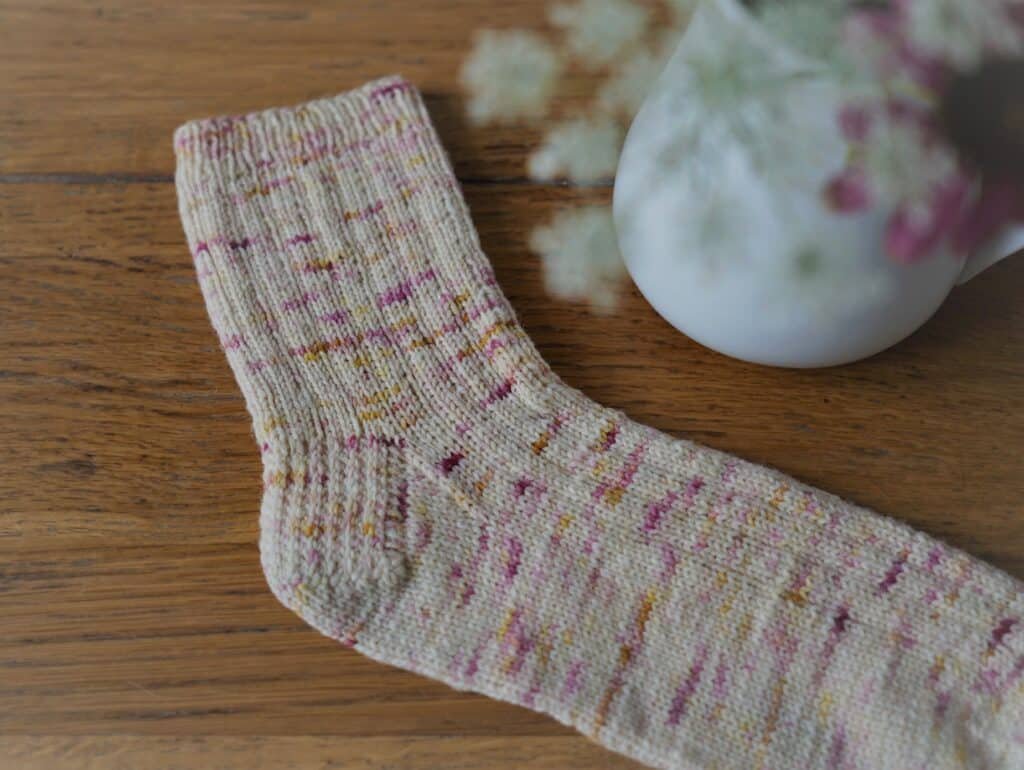 close up of a colorful hand knitted sock on a wooden table next to a vase filled with flowers