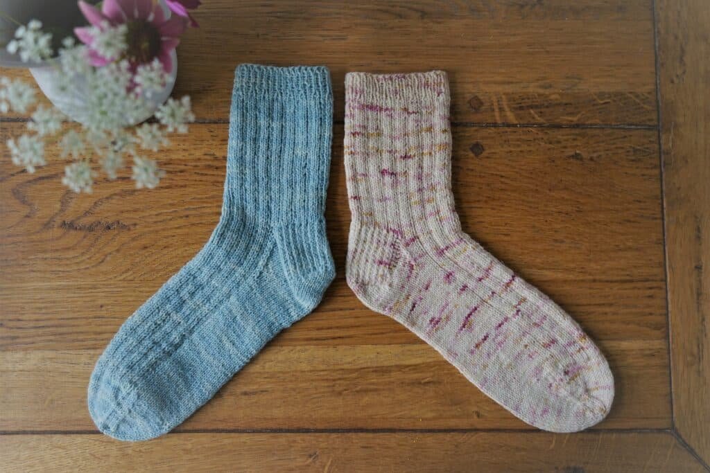 two hand knitted socks, one in light blue and one in cream with yellow and pink speckles on a wooden surface next to a vase filled with flowers
