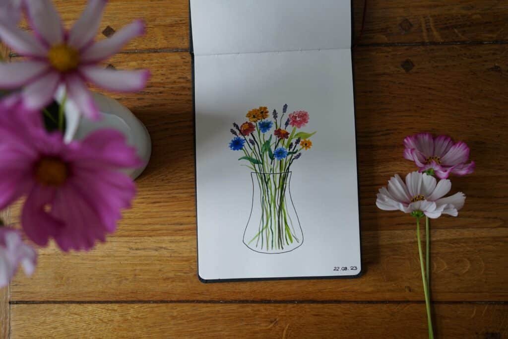 watercolor painting of wildflowers in a vase. The open sketchbook is sitting on a wooden table and there are some pink cosmos next to it.