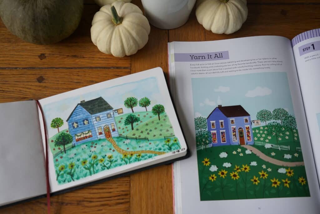 open sketchbook with a watercolor and ink painting of a blue yarn shop next to a book and some pumpkins. everything is sitting on a wooden table.