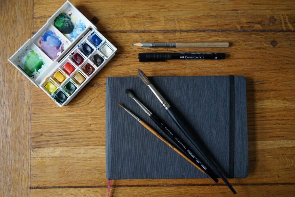 sketchbook, three pencils, an ink pen, a pen and a used watercolor palette on a wooden table