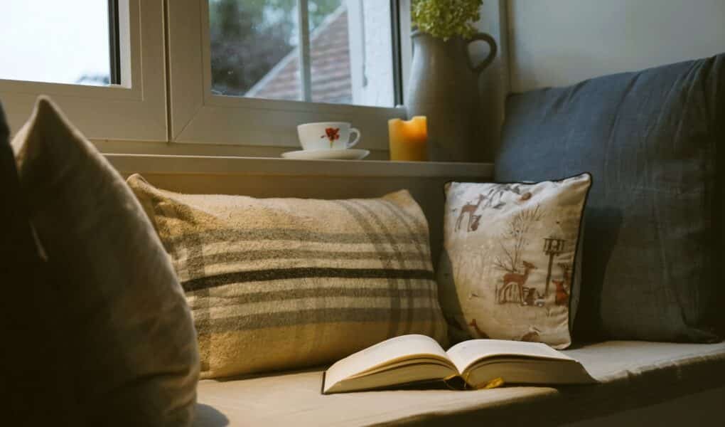 a window seat with several cozy pillows, an open book, a lit beeswax candle, a vase with a dried hydrangea and a cup of teas sitting on the window sill