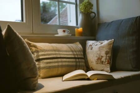 a window seat with several cozy pillows, an open book, a lit beeswax candle, a vase with a dried hydrangea and a cup of teas sitting on the window sill