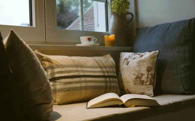 a window seat with several cozy pillows, an open book, a lit beeswax candle, a vase with a dried hydrangea and a cup of teas sitting on the window sill