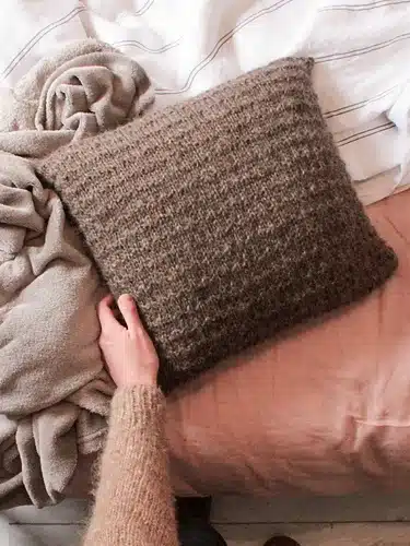 a hand touching a handknitted brown pillow next to some blankets
