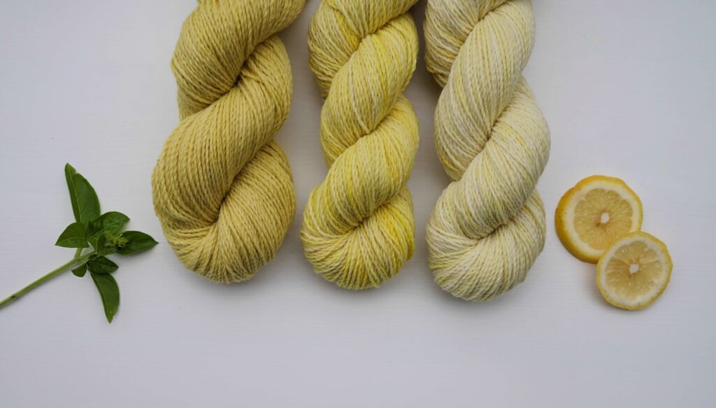 three skeins of different shades of yellow, naturally dyed yarn next to two citrus slices and a branch of mint. everything is laying on a white surface