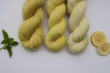 three skeins of different shades of yellow, naturally dyed yarn next to two citrus slices and a branch of mint. everything is laying on a white surface