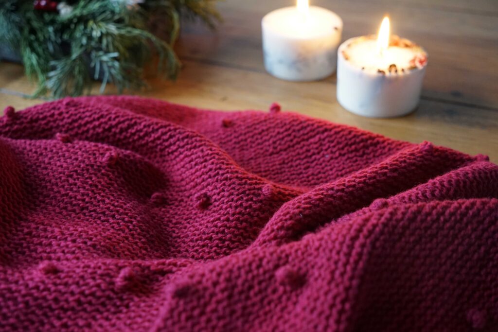 close up of a red hand knitted shawl with bobbles and garter stitch on a wooden table, two burning candles in the background