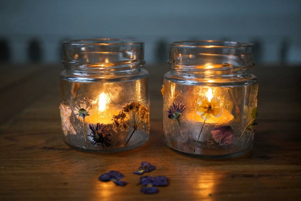 two glass jar candle holders with pressed flowers and two burning beeswax tealights in them. the candle holders are standing on a wooden table. next to them lay 3 pressed violet flowers.