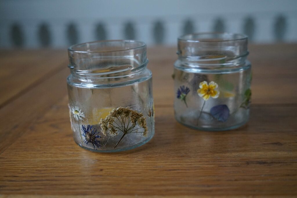two glass jars with different pressed flowers applied to them. the jars are sitting on a wooden table.